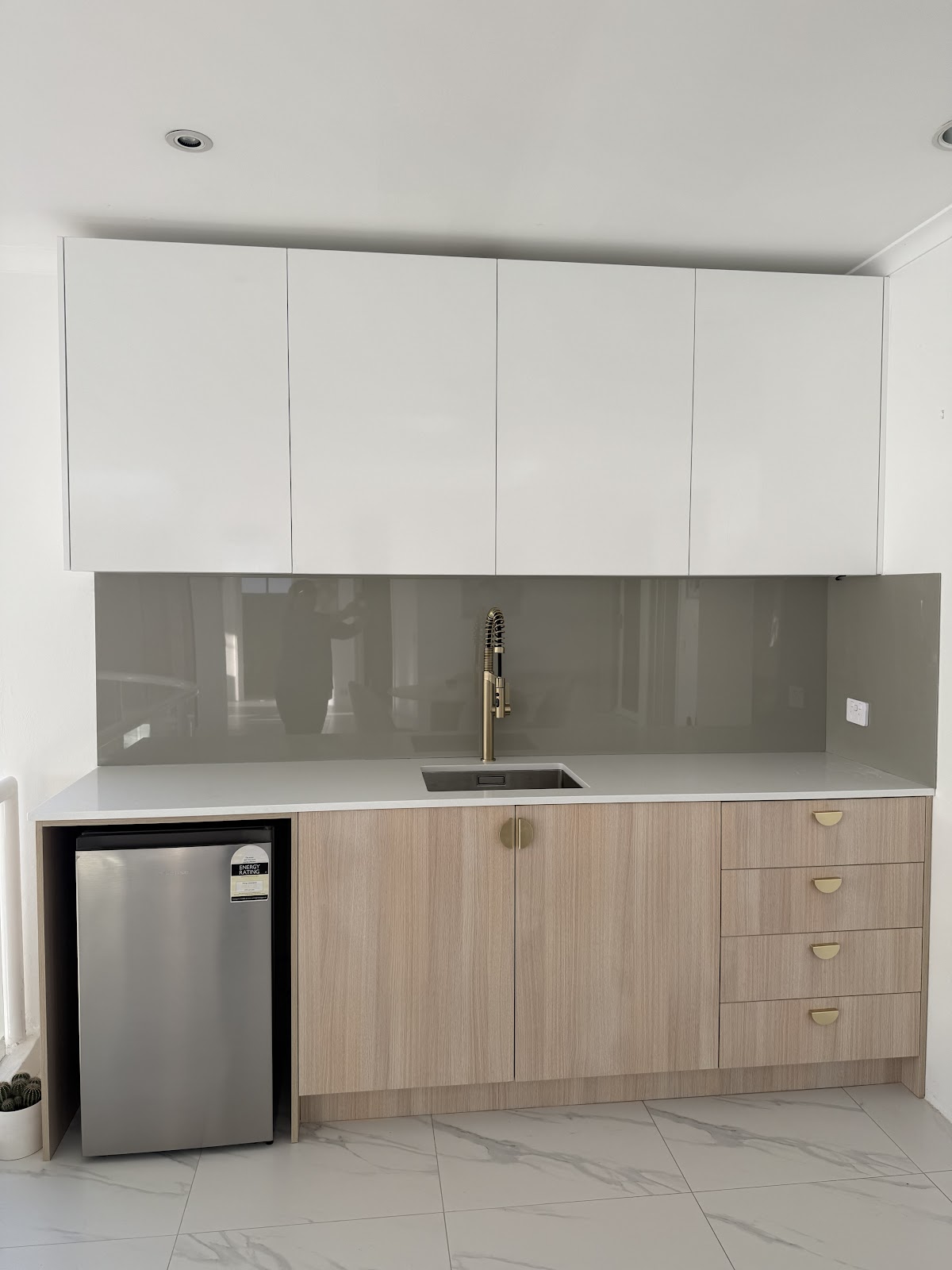 A kitchenette with white overhead cupboards, timber-look base cabinets with brass pulls and a bar fridge nook